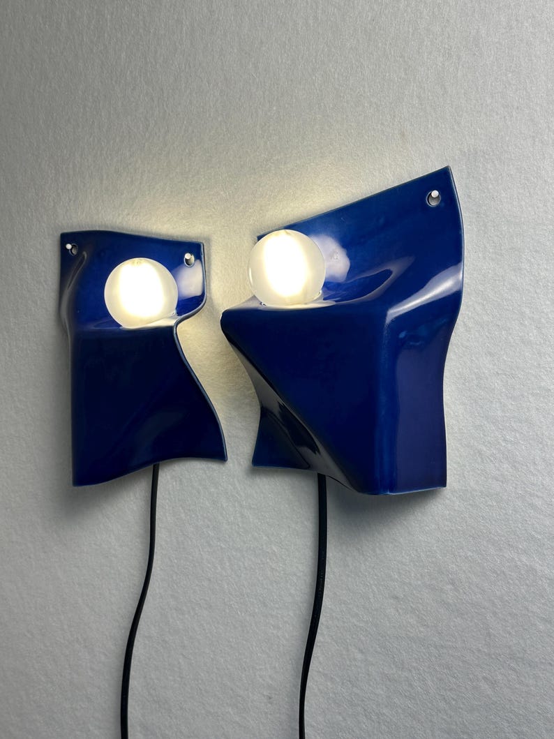 May include: Two wall-mounted lamps featuring glossy, deep blue ceramic shades with irregular, folded forms. Each lamp includes a single, spherical, glowing white light bulb nestled within the ceramic structure and a black power cord extending downward.