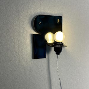 May include: A wall-mounted lamp featuring a glossy, dark blue, abstract-shaped ceramic backplate. A single, round, glowing white light bulb is attached to a black socket extending from the right side, with a thin, clear power cord hanging downward.