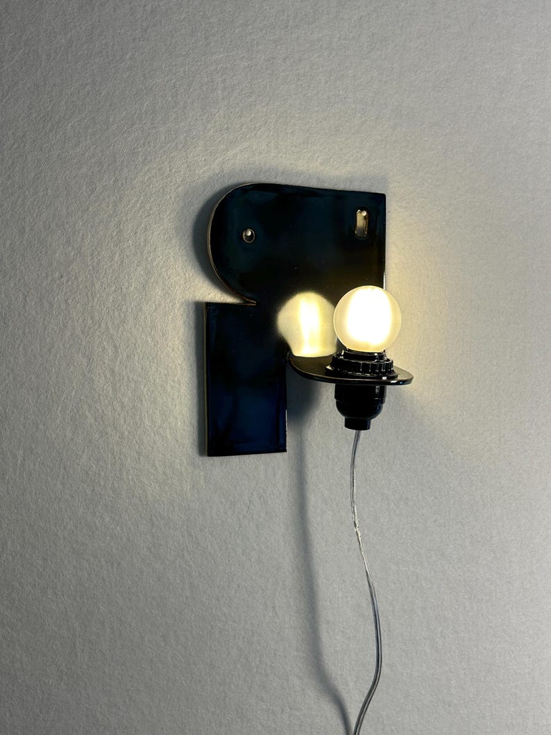 May include: A wall-mounted lamp featuring a glossy, dark blue, abstract-shaped ceramic backplate. A single, round, glowing white light bulb is attached to a black socket extending from the right side, with a thin, clear power cord hanging downward.