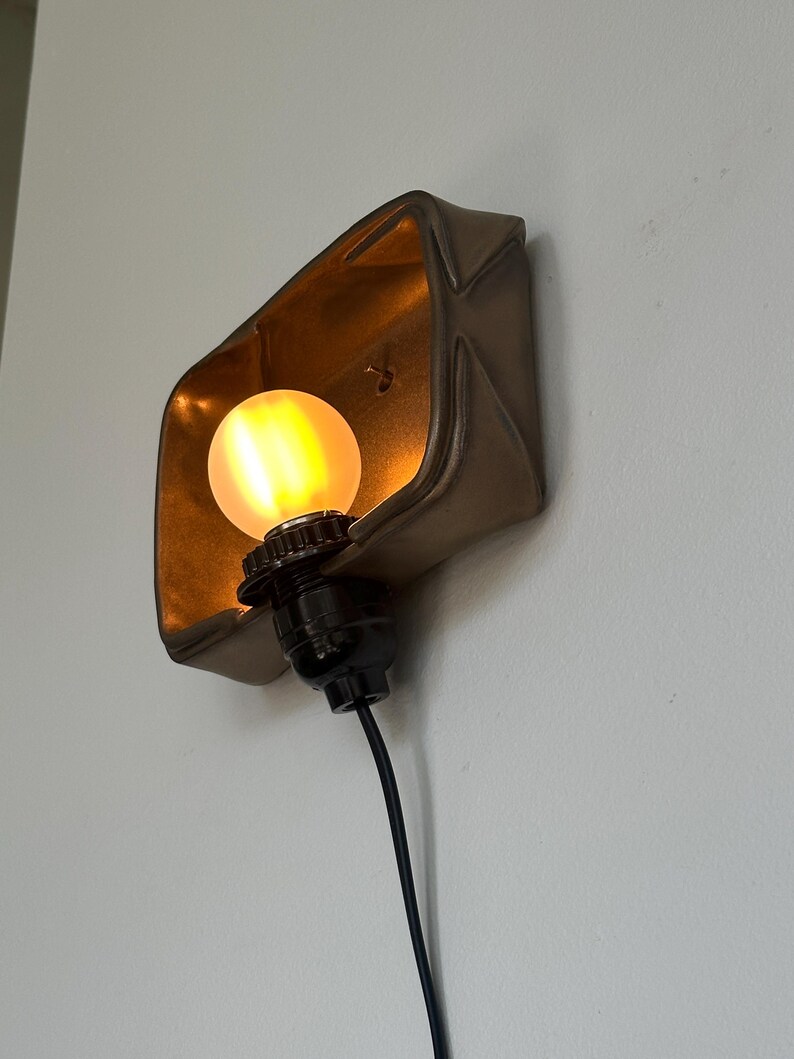 May include: A bronze-colored wall sconce with a warm, glowing light bulb. The sconce has a geometric, folded design, and the bulb is encased within. A black electrical cord extends from the fixture.
