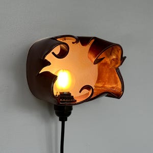 May include: A brown leather wall sconce with a decorative cutout design. The sconce houses a lit lightbulb, casting a warm glow. A black cord extends from the base, ready for plugging in. The fixture is mounted on a light gray wall.