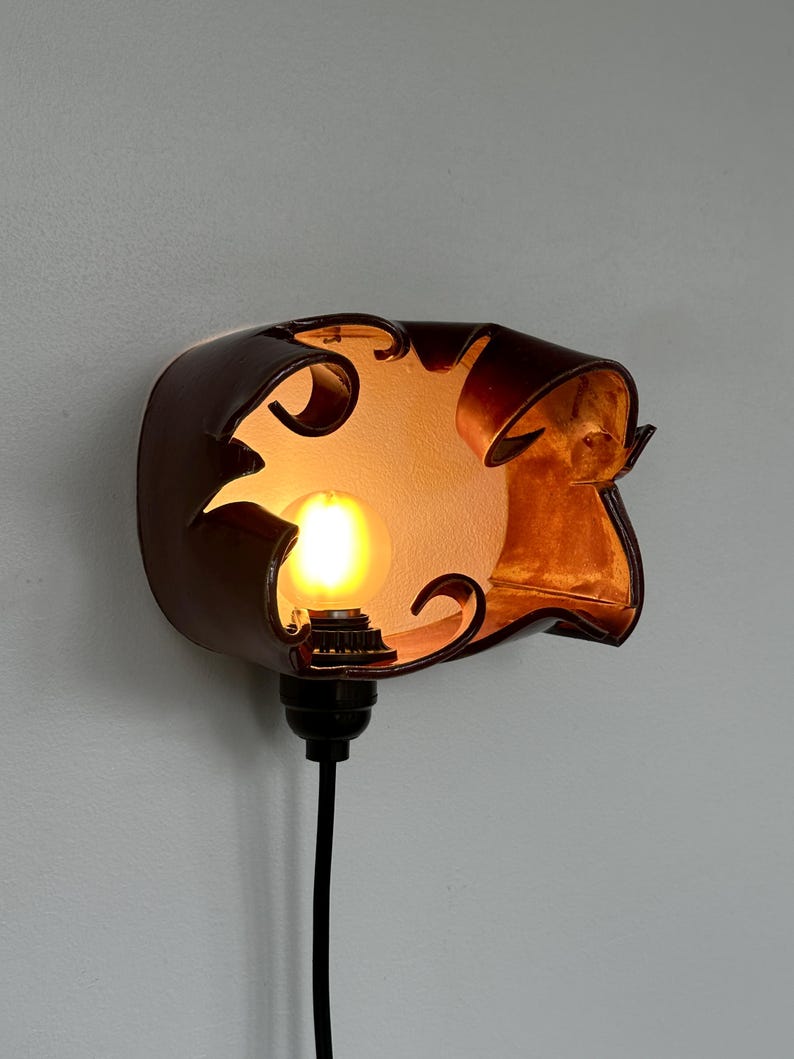 May include: A brown leather wall sconce with a decorative cutout design. The sconce houses a lit lightbulb, casting a warm glow. A black cord extends from the base, ready for plugging in. The fixture is mounted on a light gray wall.
