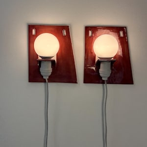 Puede incluir: Un par de apliques de pared con placas traseras de cer&aacute;mica de color rojo oscuro, brillantes y de forma trapezoidal. Cada l&aacute;mpara incluye una bombilla esf&eacute;rica blanca, un casquillo blanco y un cable de alimentaci&oacute;n blanco.