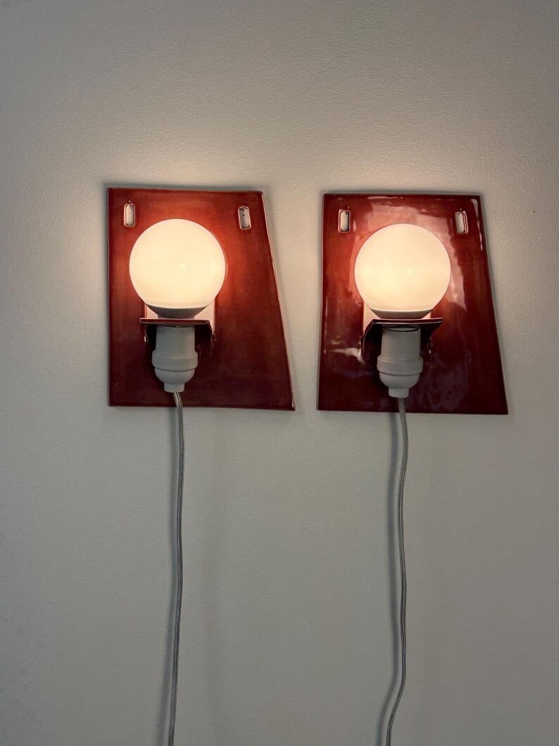 Puede incluir: Un par de apliques de pared con placas traseras de cer&aacute;mica de color rojo oscuro, brillantes y de forma trapezoidal. Cada l&aacute;mpara incluye una bombilla esf&eacute;rica blanca, un casquillo blanco y un cable de alimentaci&oacute;n blanco.
