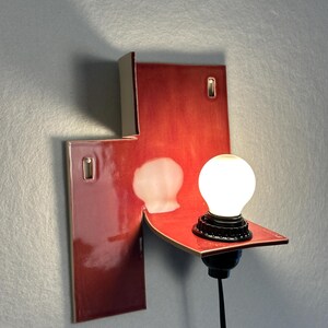 Pode incluir: Arandela de parede moderna com acabamento vermelho brilhante. A lumin&aacute;ria apresenta um design geom&eacute;trico &uacute;nico com uma pequena prateleira que suporta uma l&acirc;mpada redonda e branca. Montada em uma parede cinza.