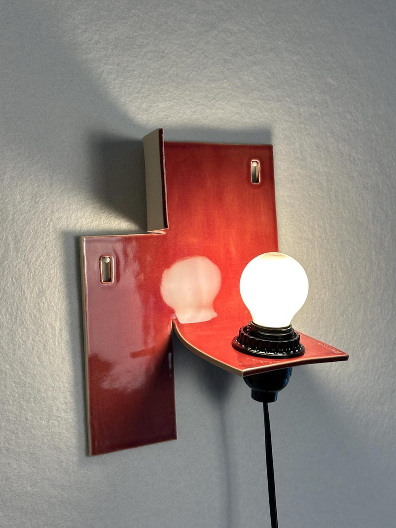 Pode incluir: Arandela de parede moderna com acabamento vermelho brilhante. A lumin&aacute;ria apresenta um design geom&eacute;trico &uacute;nico com uma pequena prateleira que suporta uma l&acirc;mpada redonda e branca. Montada em uma parede cinza.