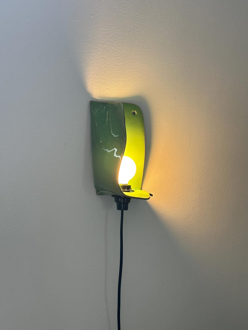 Pu&ograve; includere: Una lampada da parete con un paralume in ceramica verde lucido e curvo con una finitura liscia. L'apparecchio ospita una singola lampadina rotonda a vista che emette una luce calda, proiettando luce sulla parete. Un cavo nero scende verso il basso.