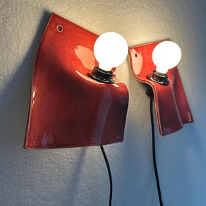 Puede incluir: Dos l&aacute;mparas de pared con pantallas de cer&aacute;mica roja brillante de formas curvas y superficie ondulada. Cada l&aacute;mpara incluye una bombilla redonda de luz blanca en un casquillo negro y un cable de alimentaci&oacute;n negro hacia abajo.