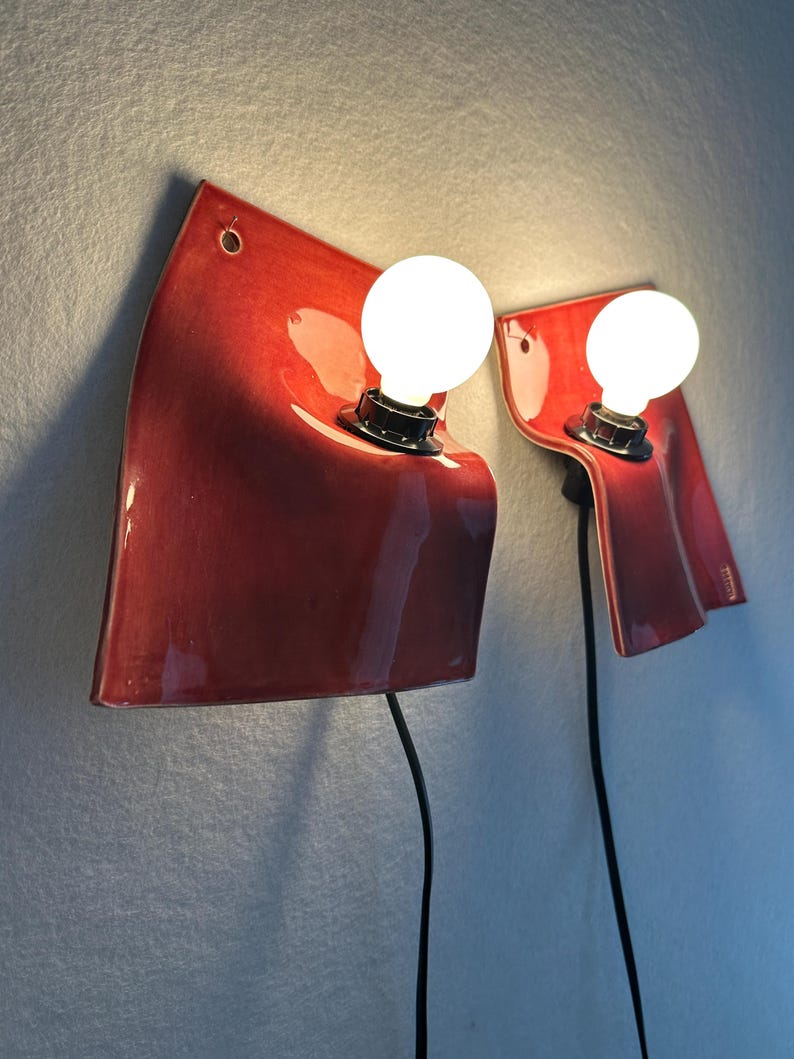 Puede incluir: Dos l&aacute;mparas de pared con pantallas de cer&aacute;mica roja brillante de formas curvas y superficie ondulada. Cada l&aacute;mpara incluye una bombilla redonda de luz blanca en un casquillo negro y un cable de alimentaci&oacute;n negro hacia abajo.