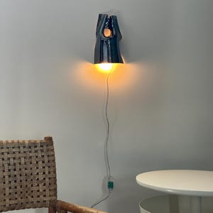 May include: A wall-mounted ceramic light fitting with a glossy dark blue finish and an organic, elongated shape. The fitting emits a warm, glowing light from its base and includes a white power cord with an integrated switch hanging downwards.