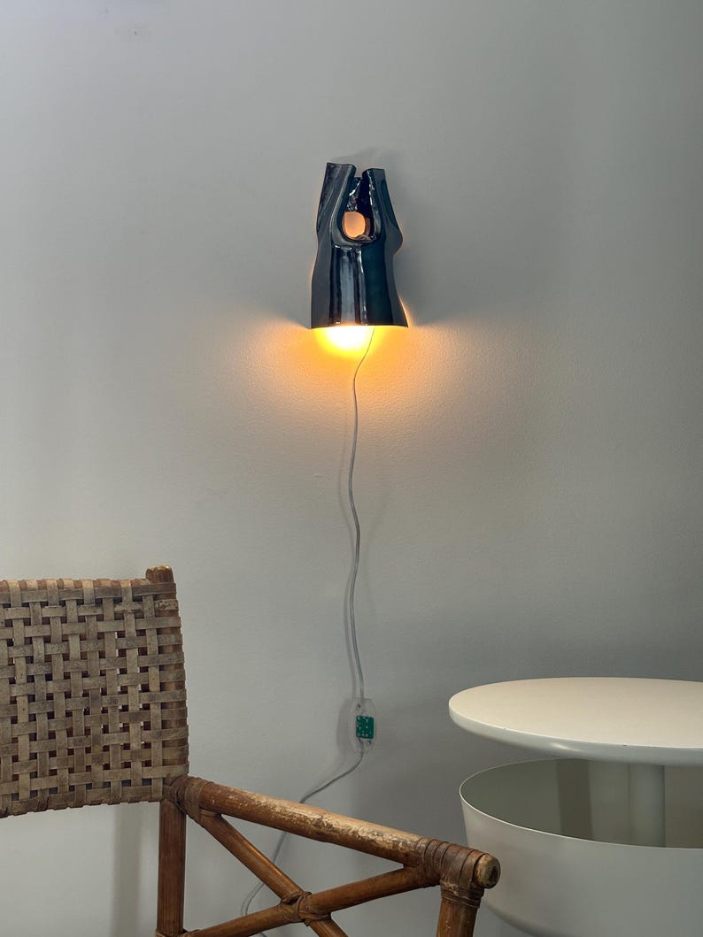 May include: A wall-mounted ceramic light fitting with a glossy dark blue finish and an organic, elongated shape. The fitting emits a warm, glowing light from its base and includes a white power cord with an integrated switch hanging downwards.