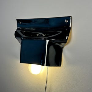 May include: A glossy black ceramic wall sconce with a curved, organic shape. The fixture features two mounting holes at the top and a central opening that holds a single round, glowing light bulb, with a thin power cord extending downward.