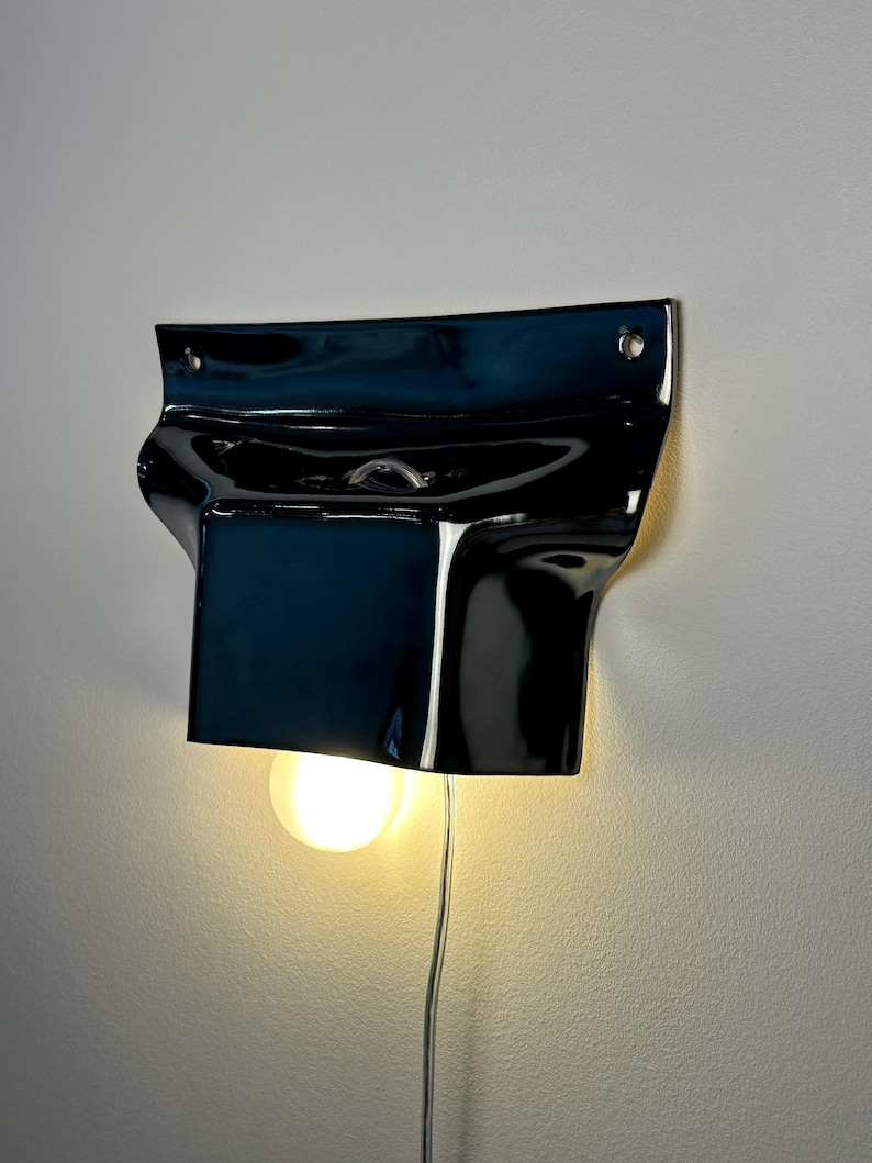May include: A glossy black ceramic wall sconce with a curved, organic shape. The fixture features two mounting holes at the top and a central opening that holds a single round, glowing light bulb, with a thin power cord extending downward.