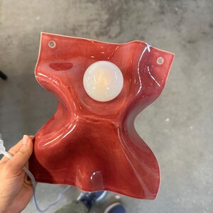 Puede incluir: Una l&aacute;mpara de pared con una pantalla de cer&aacute;mica ondulada de color rojo intenso brillante y forma fluida y org&aacute;nica. El aplique incluye una bombilla central circular de cristal blanco esmerilado y dos orificios de montaje en las esquinas.
