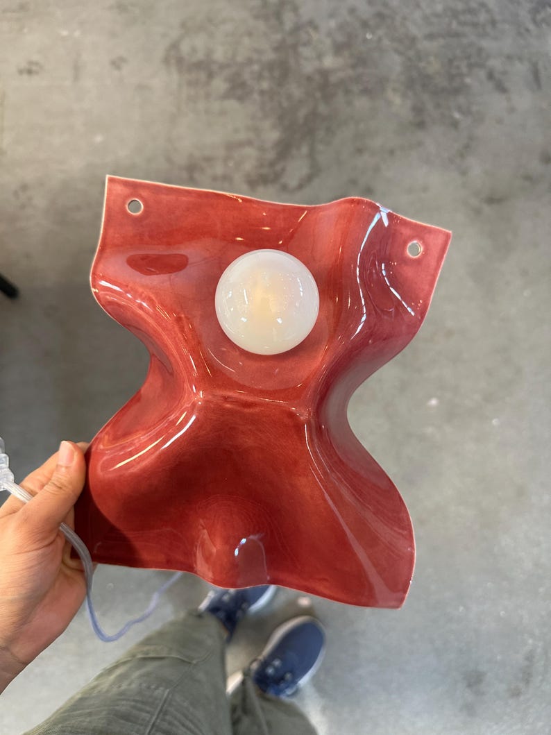 Puede incluir: Una l&aacute;mpara de pared con una pantalla de cer&aacute;mica ondulada de color rojo intenso brillante y forma fluida y org&aacute;nica. El aplique incluye una bombilla central circular de cristal blanco esmerilado y dos orificios de montaje en las esquinas.