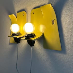 May include: A pair of wall-mounted sconces featuring bright yellow, curved ceramic backplates with integrated black light sockets and glowing round white bulbs. Each fixture includes a clear power cord extending downward from the base.
