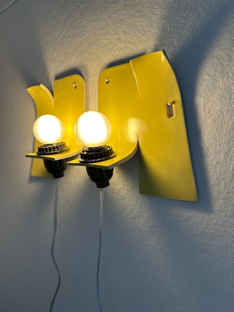 May include: A pair of wall-mounted sconces featuring bright yellow, curved ceramic backplates with integrated black light sockets and glowing round white bulbs. Each fixture includes a clear power cord extending downward from the base.