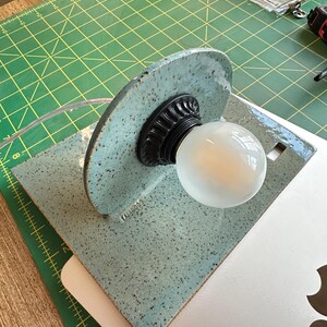 May include: A light blue ceramic lamp with a speckled finish and a semi-circular backplate. The lamp features a black fluted socket holding a round white light bulb. The word "TAJAGOOD" is embossed on the lower left side of the ceramic base.