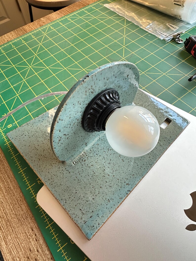 May include: A light blue ceramic lamp with a speckled finish and a semi-circular backplate. The lamp features a black fluted socket holding a round white light bulb. The word "TAJAGOOD" is embossed on the lower left side of the ceramic base.