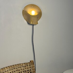 May include: A wall-mounted lamp with a smooth, cream-colored, shell-shaped shade that curves outward. The lamp emits a warm, glowing light from a central bulb and features a thin, black electrical cord extending downward from the base of the fixture.