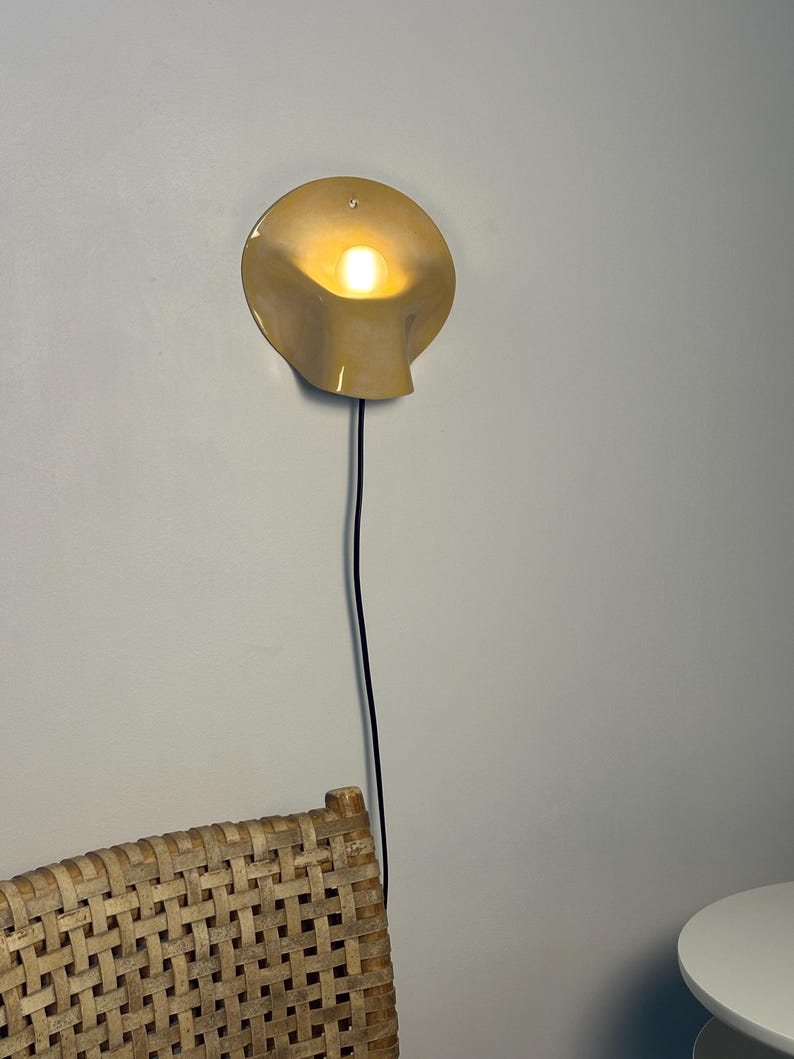 May include: A wall-mounted lamp with a smooth, cream-colored, shell-shaped shade that curves outward. The lamp emits a warm, glowing light from a central bulb and features a thin, black electrical cord extending downward from the base of the fixture.