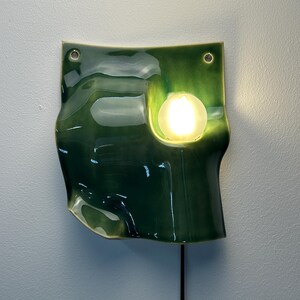 May include: A wall-mounted ceramic lamp with a glossy, deep green glaze and an irregular, wavy shape. The fixture features two circular holes at the top for mounting and a single, round, glowing light bulb set into a recessed area on the right side.