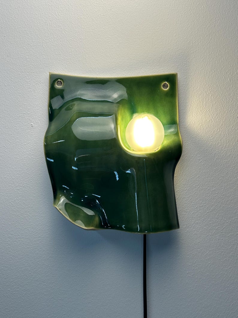 May include: A wall-mounted ceramic lamp with a glossy, deep green glaze and an irregular, wavy shape. The fixture features two circular holes at the top for mounting and a single, round, glowing light bulb set into a recessed area on the right side.