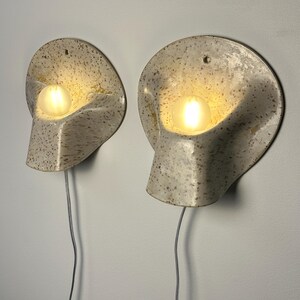 May include: A pair of wall-mounted sconces featuring a speckled, off-white ceramic shell with a curved, organic shape. Each fixture holds a single glowing, round light bulb and includes a thin, light-coloured electrical cord extending downwards.