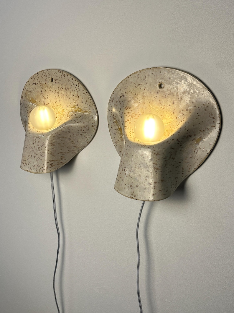 May include: A pair of wall-mounted sconces featuring a speckled, off-white ceramic shell with a curved, organic shape. Each fixture holds a single glowing, round light bulb and includes a thin, light-coloured electrical cord extending downwards.