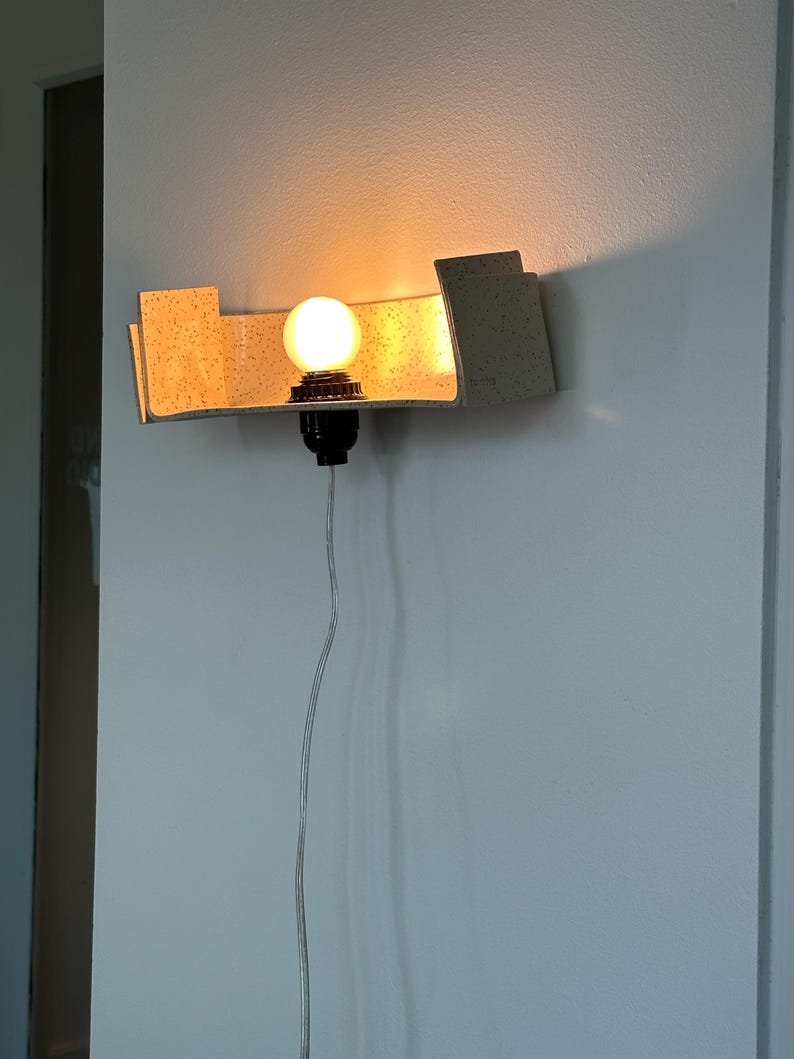 May include: A modern wall sconce with a warm, glowing light bulb. The sconce has a beige, speckled shade and a black bulb socket. The light is mounted on a white wall, with a visible power cord.