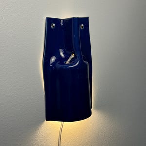 Puede incluir: Un aplique de pared de cer&aacute;mica azul oscuro brillante con una forma escult&oacute;rica curva. La pieza cuenta con dos peque&ntilde;os orificios de montaje circulares en la parte superior y una abertura central por donde sale un cable de luz.
