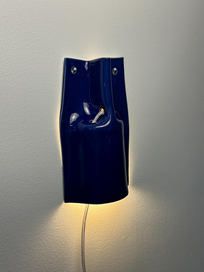 Puede incluir: Un aplique de pared de cer&aacute;mica azul oscuro brillante con una forma escult&oacute;rica curva. La pieza cuenta con dos peque&ntilde;os orificios de montaje circulares en la parte superior y una abertura central por donde sale un cable de luz.