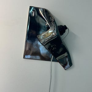Peut inclure: Une applique murale compos&eacute;e d'un abat-jour en c&eacute;ramique m&eacute;tallique sombre et r&eacute;fl&eacute;chissante, pr&eacute;sentant une forme irr&eacute;guli&egrave;re et pli&eacute;e. Le luminaire comprend une douille noire et un fin cordon &eacute;lectrique clair descendant de la base en c&eacute;ramique.