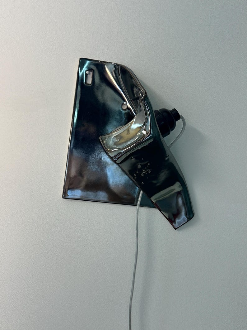 Peut inclure: Une applique murale compos&eacute;e d'un abat-jour en c&eacute;ramique m&eacute;tallique sombre et r&eacute;fl&eacute;chissante, pr&eacute;sentant une forme irr&eacute;guli&egrave;re et pli&eacute;e. Le luminaire comprend une douille noire et un fin cordon &eacute;lectrique clair descendant de la base en c&eacute;ramique.
