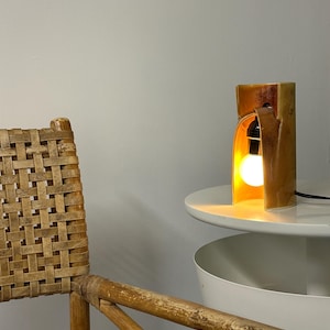 Puede incluir: Una l&aacute;mpara de mesa de cer&aacute;mica con un esmalte brillante en tonos &aacute;mbar y una estructura curva en forma de arco. La l&aacute;mpara cuenta con un casquillo negro que sostiene una bombilla redonda brillante, emitiendo una luz c&aacute;lida desde el interior.