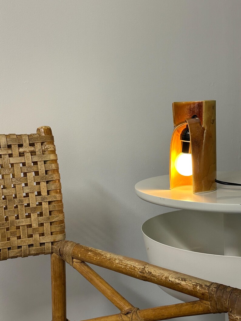 Puede incluir: Una l&aacute;mpara de mesa de cer&aacute;mica con un esmalte brillante en tonos &aacute;mbar y una estructura curva en forma de arco. La l&aacute;mpara cuenta con un casquillo negro que sostiene una bombilla redonda brillante, emitiendo una luz c&aacute;lida desde el interior.
