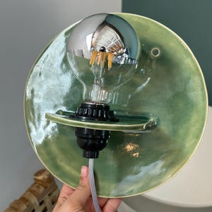 May include: A wall-mounted light fixture featuring a circular, sage green glazed ceramic backplate with a glossy finish. A black socket holds a clear glass light bulb with a reflective silver top half and visible internal orange filaments.