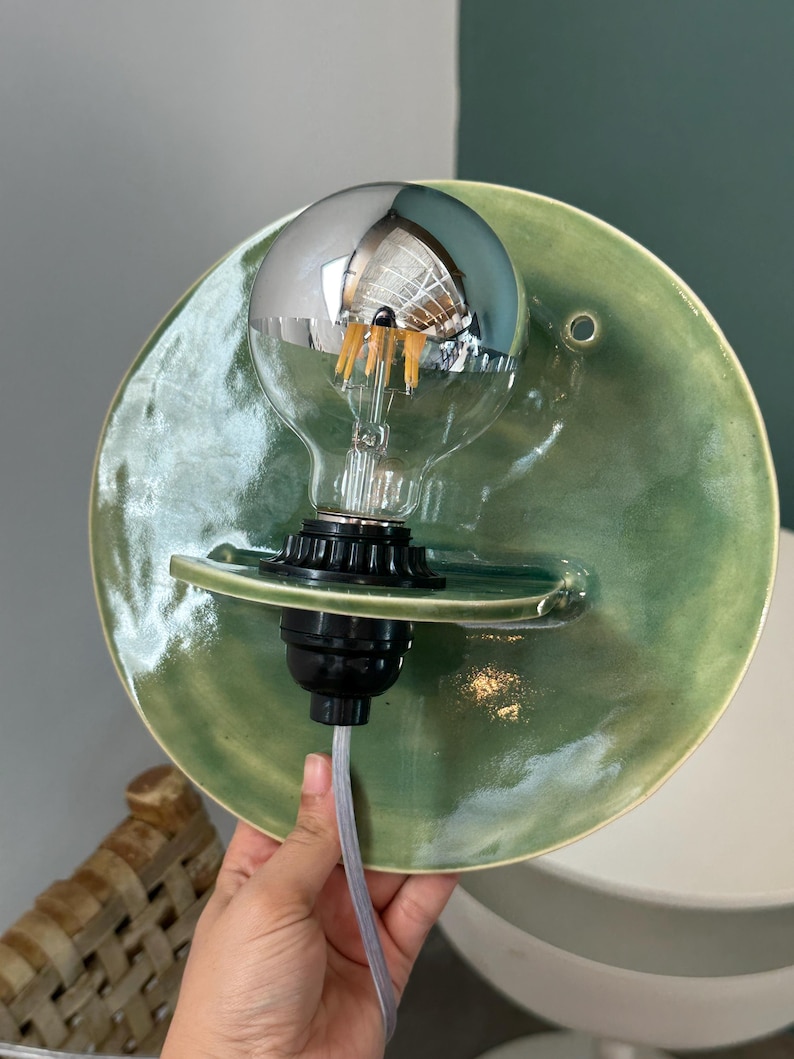 May include: A wall-mounted light fixture featuring a circular, sage green glazed ceramic backplate with a glossy finish. A black socket holds a clear glass light bulb with a reflective silver top half and visible internal orange filaments.