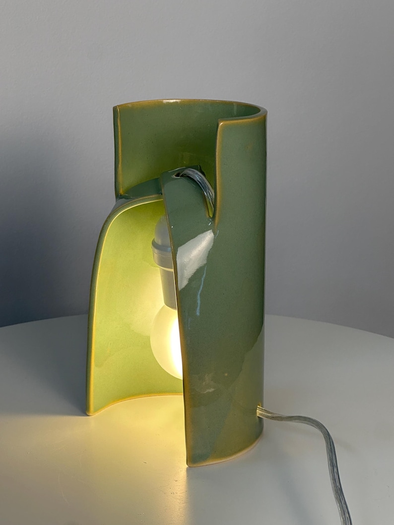 May include: A cylindrical ceramic table lamp with a glossy sage green glaze. The lamp features a curved, open-front design that reveals a white light bulb socket and a glowing bulb inside. A thin, light-colored electrical cord extends from the base.