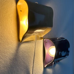 May include: Two wall-mounted ceramic sconces with a curved, organic shape. The top sconce is a glossy yellow, while the bottom sconce features a deep purple interior and a dark exterior. Each lamp emits a warm, glowing light from a central bulb.