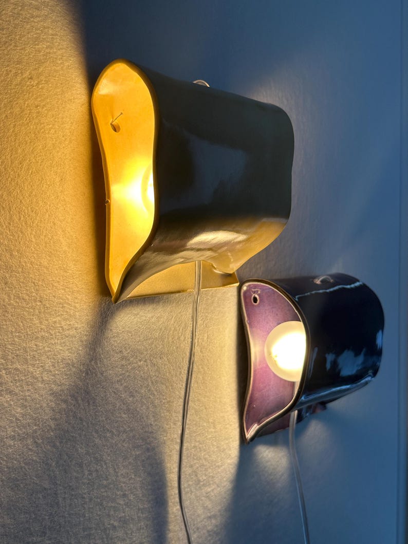 May include: Two wall-mounted ceramic sconces with a curved, organic shape. The top sconce is a glossy yellow, while the bottom sconce features a deep purple interior and a dark exterior. Each lamp emits a warm, glowing light from a central bulb.