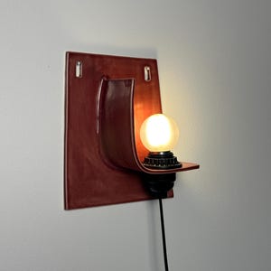 May include: A wall-mounted sconce featuring a glossy, deep red ceramic backplate with a curved shelf design. A black socket holds a glowing, frosted spherical light bulb. A thin black power cord extends downwards from the base of the light fixture.