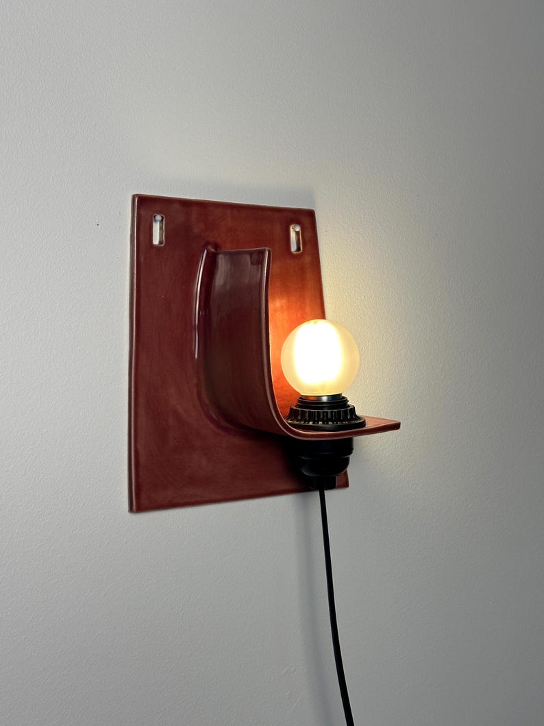 May include: A wall-mounted sconce featuring a glossy, deep red ceramic backplate with a curved shelf design. A black socket holds a glowing, frosted spherical light bulb. A thin black power cord extends downward from the base of the light fixture.