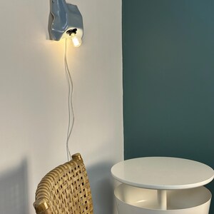 May include: A wall-mounted light fixture with a pale blue, organic, wavy ceramic shade and a single exposed light bulb. The fixture is connected to a thin white power cord that hangs vertically down the wall from the base of the lamp.