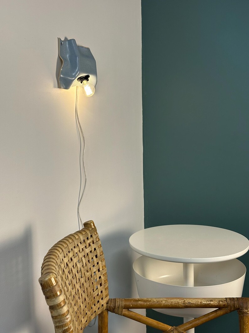 May include: A wall-mounted light fixture with a pale blue, organic, wavy ceramic shade and a single exposed light bulb. The fixture is connected to a thin white power cord that hangs vertically down the wall from the base of the lamp.