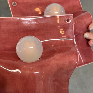 May include: Two wall-mounted light fixtures featuring a glossy, dusty rose-colored ceramic base with a wavy, organic shape. Each fixture includes a single, spherical, frosted white glass globe bulb centered on the ceramic surface and two small holes.