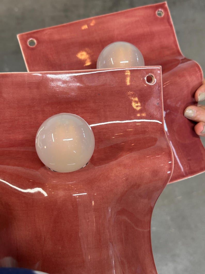 May include: Two wall-mounted light fixtures featuring a glossy, dusty rose-colored ceramic base with a wavy, organic shape. Each fixture includes a single, spherical, frosted white glass globe bulb centered on the ceramic surface and two small holes.