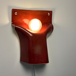 Pode incluir: Um candeeiro de parede com um corpo de cer&acirc;mica vermelho-escuro brilhante com uma forma curva e org&acirc;nica. Uma l&acirc;mpada esf&eacute;rica de vidro branco est&aacute; alojada no centro, emitindo uma luz quente. O candeeiro inclui dois orif&iacute;cios de montagem.
