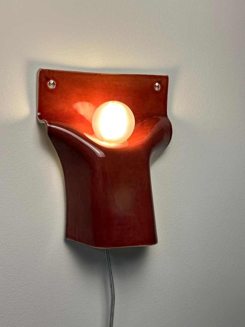 Pode incluir: Um candeeiro de parede com um corpo de cer&acirc;mica vermelho-escuro brilhante com uma forma curva e org&acirc;nica. Uma l&acirc;mpada esf&eacute;rica de vidro branco est&aacute; alojada no centro, emitindo uma luz quente. O candeeiro inclui dois orif&iacute;cios de montagem.