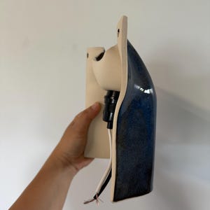 May include: A wall-mounted ceramic light fixture featuring a matte cream-colored back plate and a glossy, deep blue curved front section. The fixture includes a black socket component and exposed white and copper electrical wiring at the base.